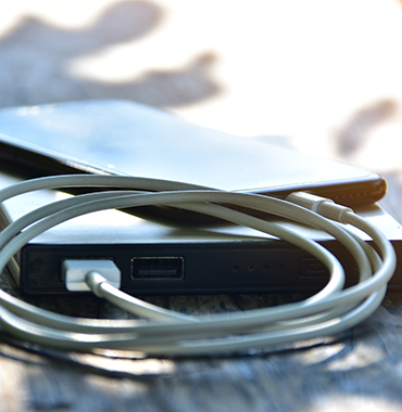 A smartphone rests on a portable power bank on a wooden surface. Two cables are wrapped around the power bank, each plugged into separate ports. The sunlight casts soft shadows on the scene, creating a bright and serene atmosphere.