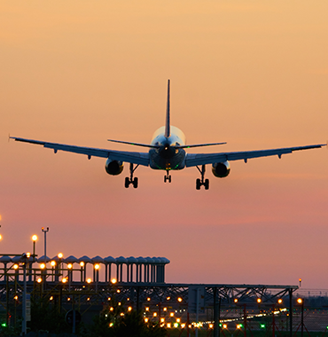 An airplane is landing at an airport during sunset. The sky is a blend of orange, pink, and purple hues, and the plane's landing gear is deployed. Airport lights and structures are visible below the descending aircraft.