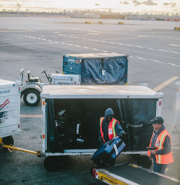 Workers wearing safety vests load luggage onto a conveyor belt from carts on an airport tarmac at sunset.