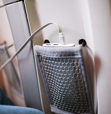 A close-up of a portable power bank charging inside a gray mesh seat pocket, attached to the back of an airplane seat. A USB cable is connected to the power bank, indicating that a device is being charged mid-flight. The airplane's interior is blurred in the background.