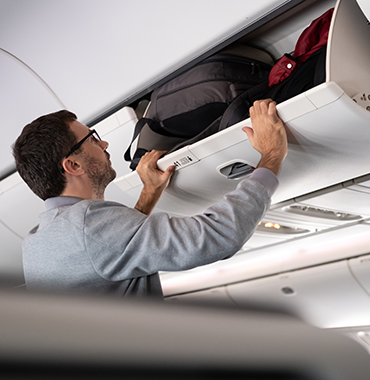 A man wearing glasses and a gray sweater is placing a black backpack into an overhead compartment on an airplane. The cabin has white walls and other overhead bins are visible.